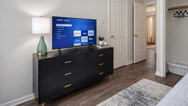 A modern bedroom with a black dresser, a flat-screen TV displaying Roku TV interface, a lamp, and a white bed. - 5 Minutes to VSU, I-75 & Restaurants! Sleeps 5 w/ 2 Full Baths- Family Friendly (Remerton)