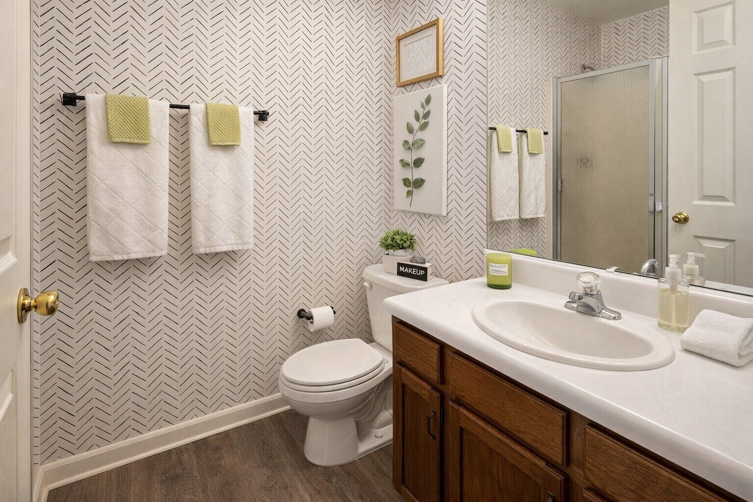 Un baño con pared de diseño en patrón de espiga, un inodoro, un lavamanos con espejo y dos toallas colgadas en un perchero.