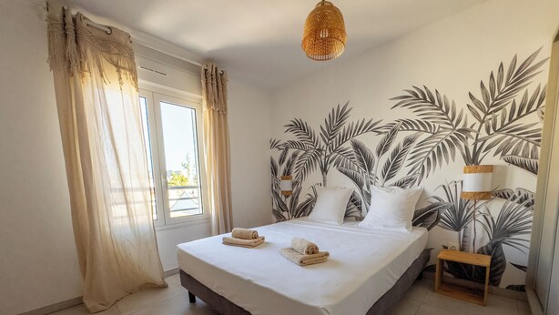 A bedroom with a bed, curtains, a window, a wall with a palm leaf mural, a small wooden nightstand, and a hanging light fixture. - EVASION SAINT LEU, APARTMENT 100 M FROM THE SEA  (Saint-Leu)