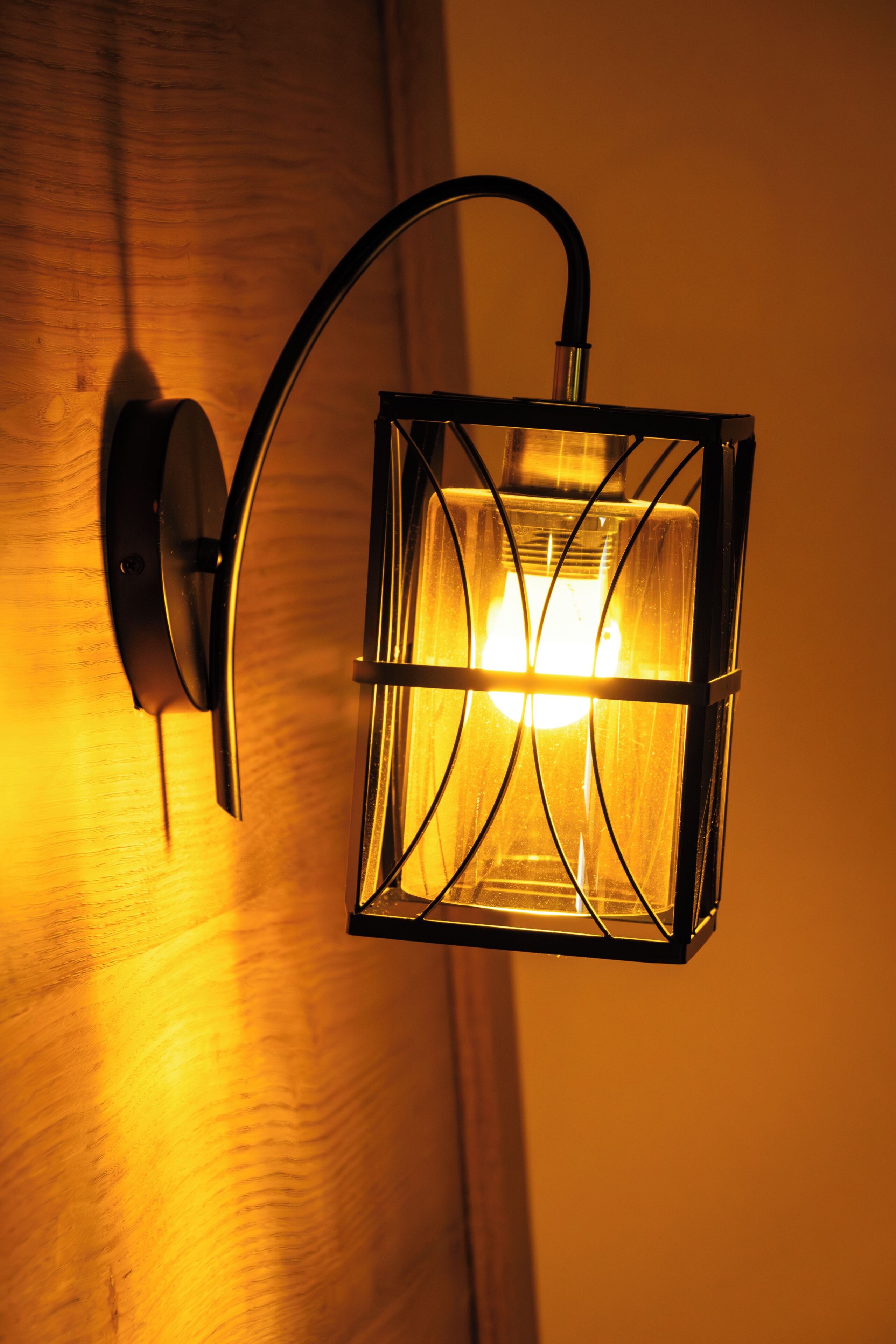 Een aan de muur bevestigde lantaarn met een gebogen arm en een vierkant glazen omhulsel, dat warm licht uitstraalt.
