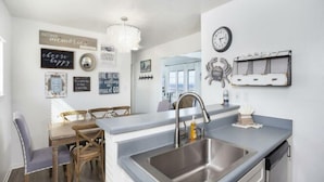 A modern kitchen with a stainless steel sink, wooden dining table, and chairs. There are wall decorations and a chandelier.