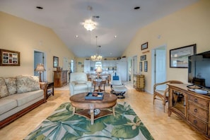 A spacious living room with a sofa, coffee table, and TV stand, a kitchen in the background, and a chandelier.