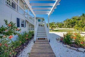 A wooden pathway leading to a white house with a porch, surrounded by a garden with red flowers and a clear blue sky.