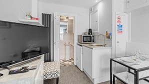A compact hotel room with a kitchenette, a flat-screen TV, a small dining table, and a bathroom visible through an open door. - Boutique Studio Apartment By My Getaways (Brighton)