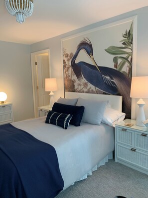 Una cama bien hecha con un edredón y almohadas azul oscuro, un gran mural de un pájaro y dos lámparas de noche sobre mesitas blancas.