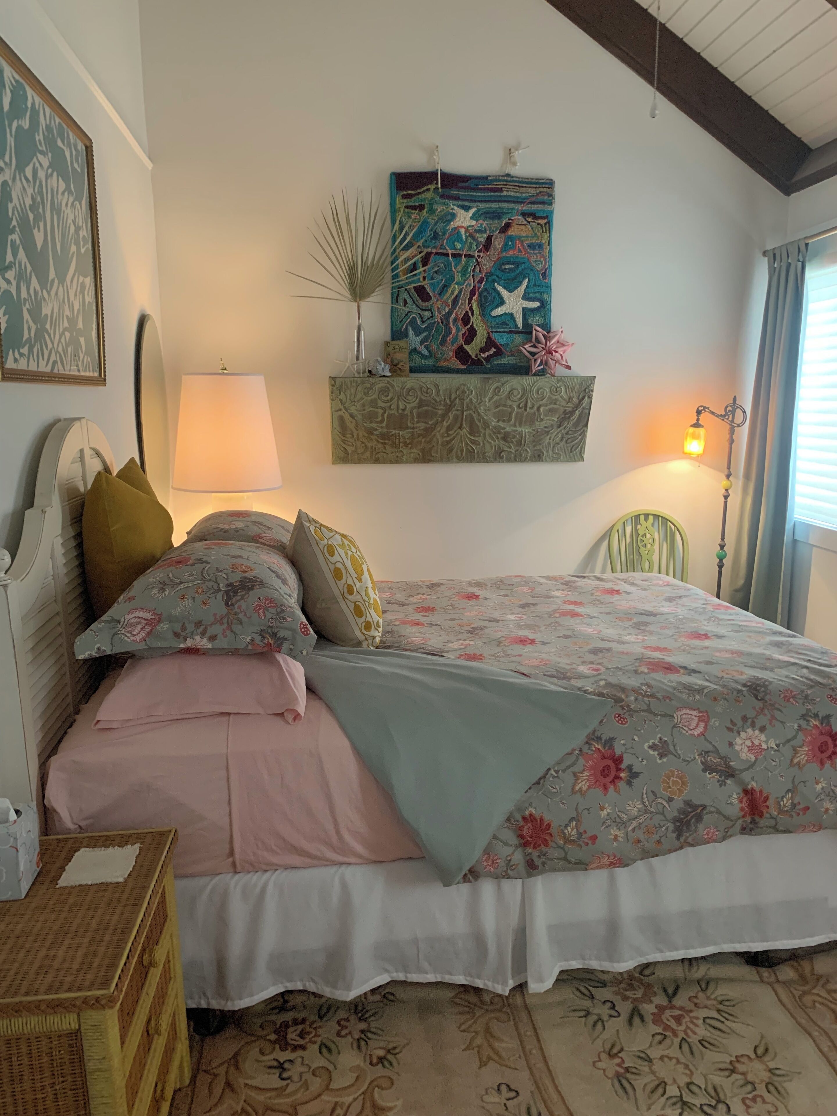 Een slaapkamer met een bed, kussens, een nachtkastje, een lamp, een decoratie aan de muur en een raam met gordijnen.