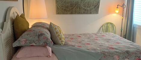 Een slaapkamer met een bed, kussens, een nachtkastje, een lamp, een decoratie aan de muur en een raam met gordijnen.