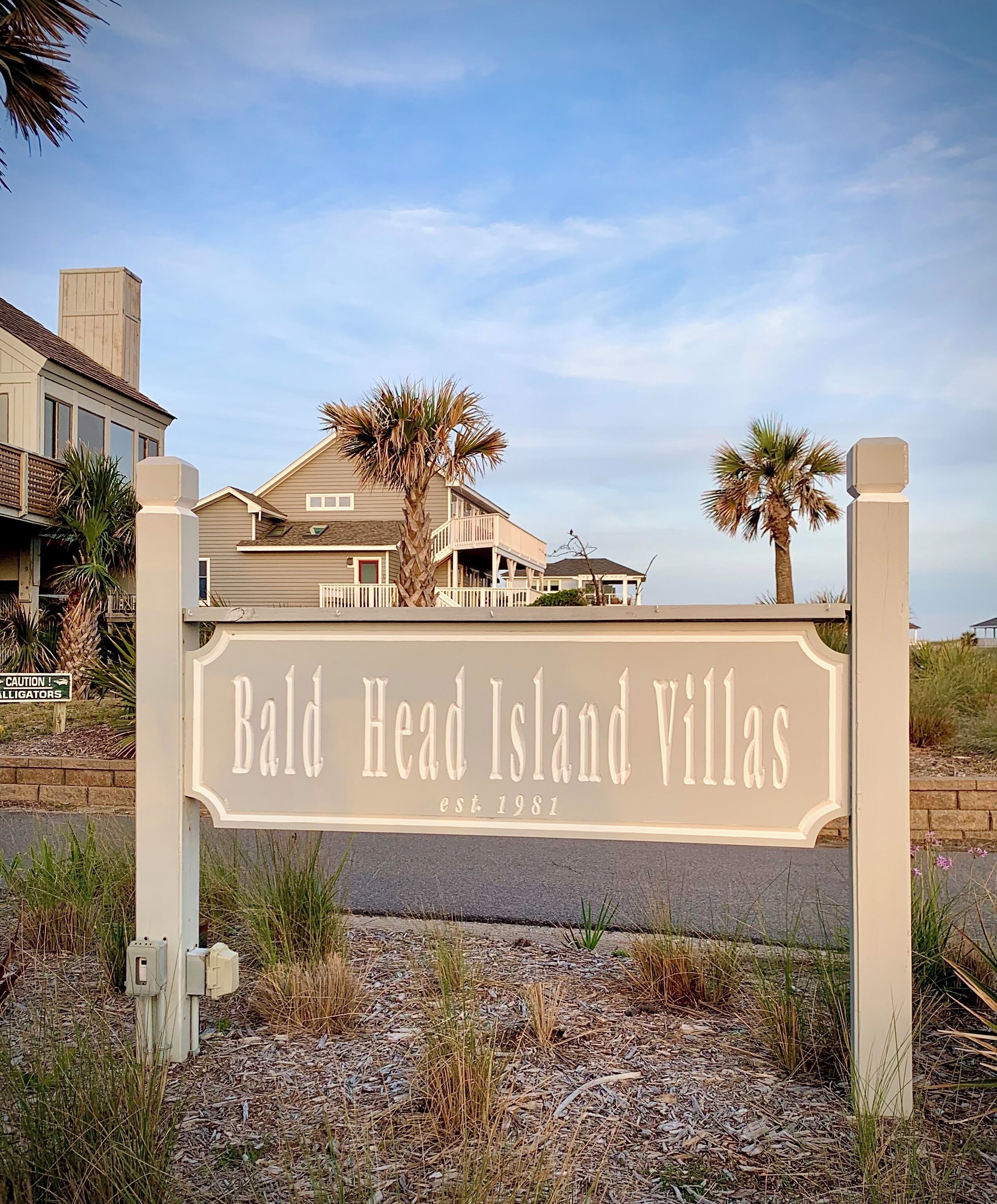 A sign for Bald Head Island Villas with a clear view of the surrounding area including a building and palm trees.