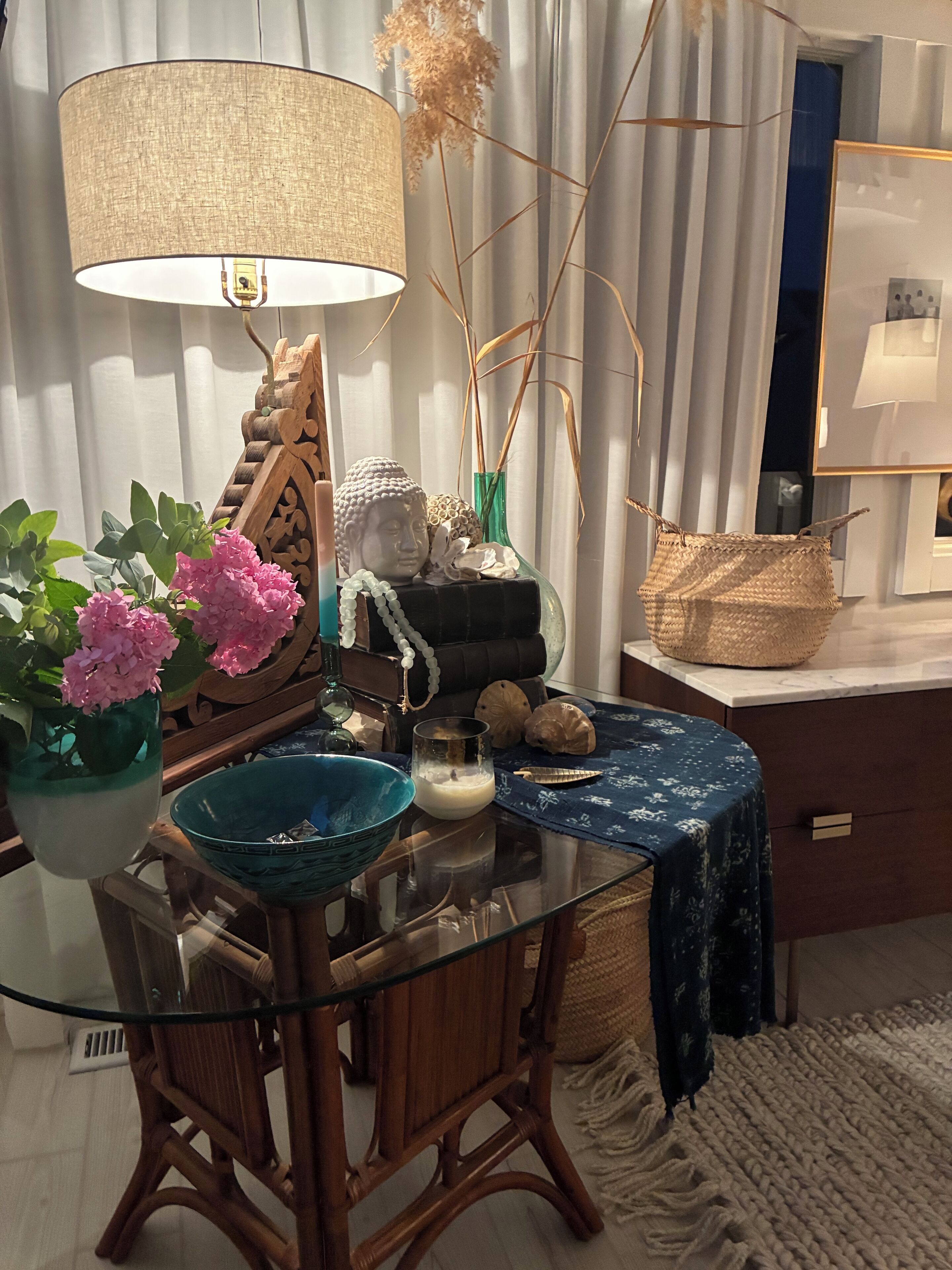 Un coin repas avec une table en verre, des chaises en bois, une nappe bleue, une tête de Bouddha décorative, un vase avec des fleurs roses, une lampe et un panier tressé.