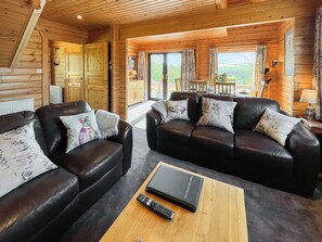 Ein gemütliches Blockhaus-Interieur mit zwei Ledersofas, einem Holztisch und einem Essbereich, der durch eine Glastür sichtbar ist.