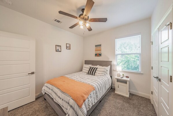 A bedroom with a bed, a nightstand, a ceiling fan, a window with blinds, and a door.