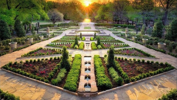 Ein symmetrisch angelegter Garten mit einem zentralen Weg, umgeben von ordentlich gestalteten Blumenbeeten und Bäumen, der zu einem Brunnen führt.
