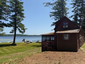A small brick cabin with a porch, situated by a lake with boats and a dock in the background. - The Little Red Cottage on Webster Lake (Franklin)