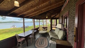 A covered outdoor area with a wooden ceiling, a table with chairs, a sofa, and a view of a lake.