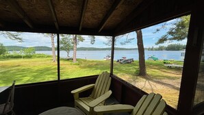 A covered porch with Adirondack chairs and a table, overlooking a grassy area with a lake and boats.