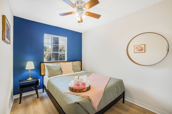 A bedroom with a bed, a bedside table with a lamp, a ceiling fan, a window with blinds, and a framed picture on the wall.