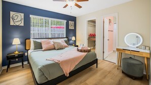 A bedroom with a bed, bedside tables, a ceiling fan, a window with a view, a wardrobe, and a vanity with a mirror.