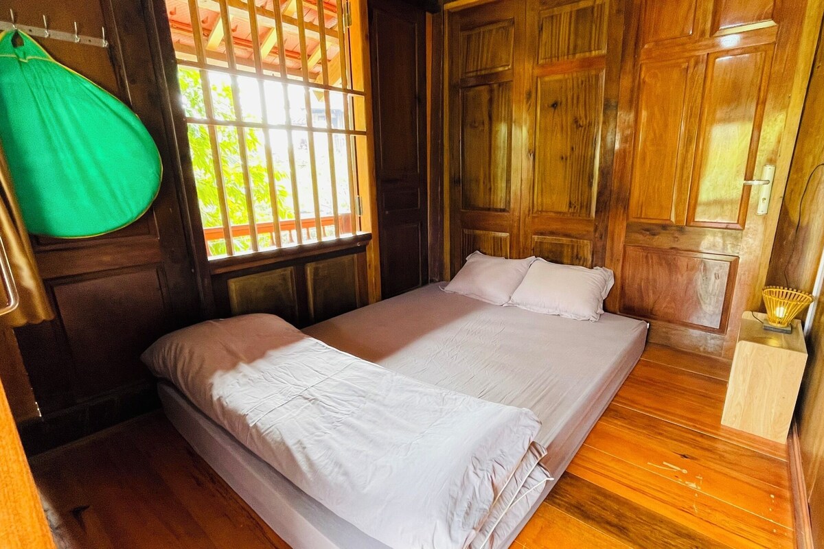A room with wooden walls and floors, a bed with white bedding, a hanging green bag, and a window with bars.