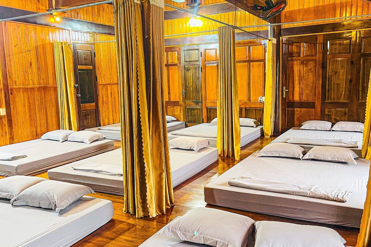 A hotel room with multiple beds arranged in a row, wooden paneling on the walls, and a ceiling fan.