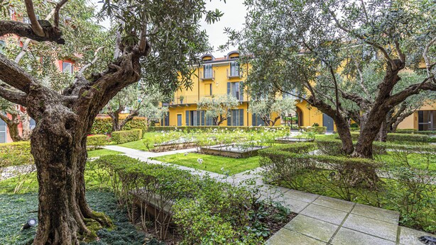 Un patio con árboles maduros, un jardín bien cuidado y un edificio amarillo con balcones.