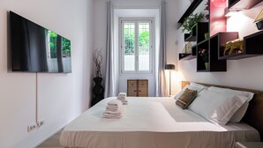 Habitación de hotel ordenada con una cama grande, un televisor de pantalla plana en la pared, un ventanal con vistas a zonas verdes y una mesita de noche con una lámpara.
