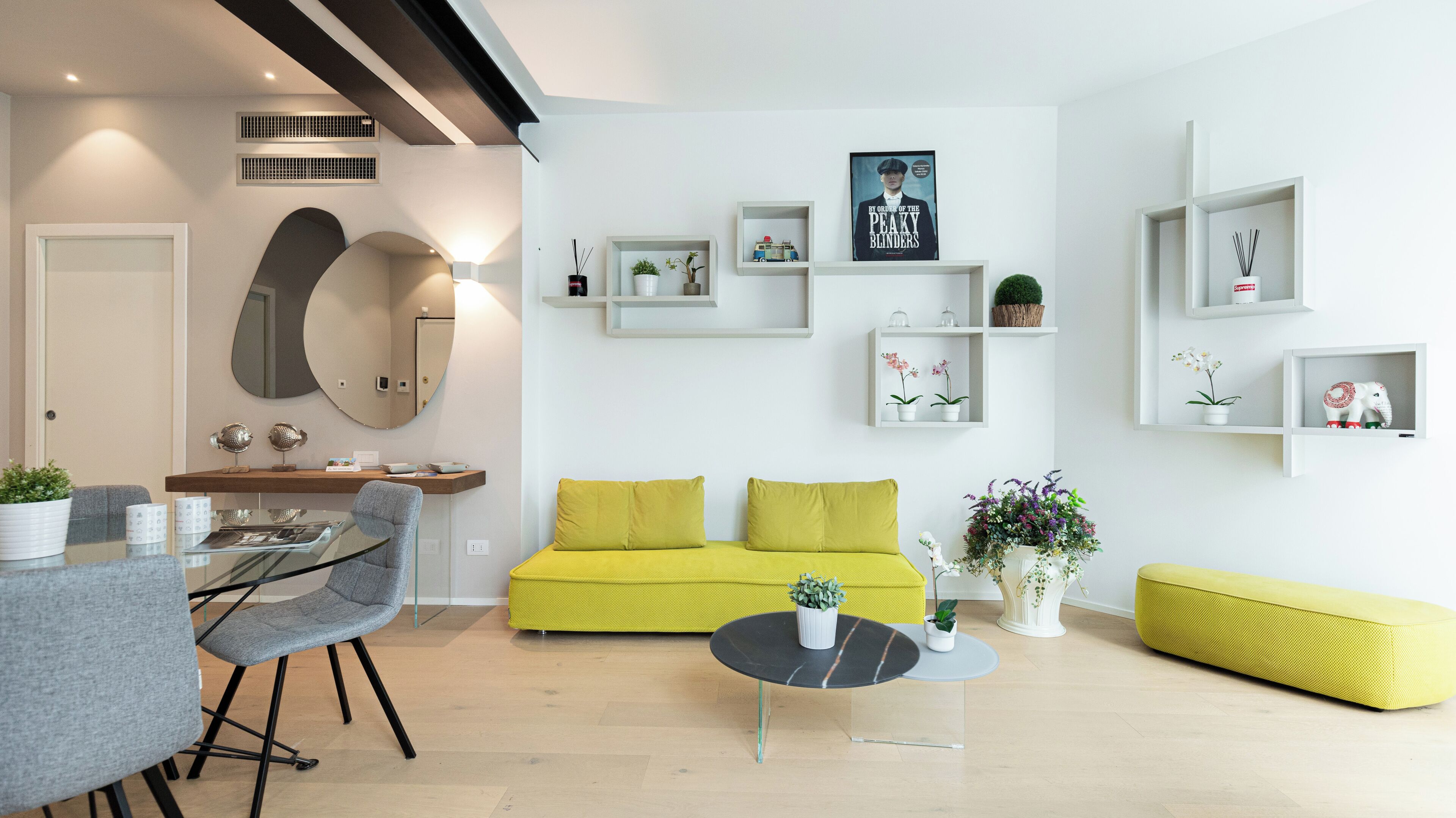 A modern living room with a yellow sofa, a glass coffee table, and a dining area with a glass table and chairs.
