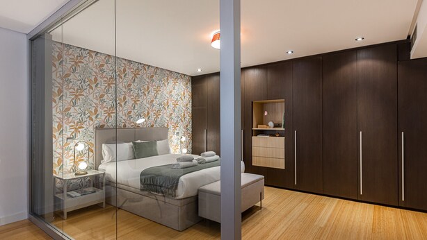 Un dormitorio moderno con separador de vidrio, una cama con almohadas, una mesita de noche y una mesita auxiliar. Se observa un armario de madera con espejo y un estante superior.