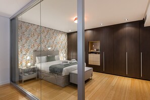 A modern bedroom with a glass partition, a bed with pillows, a bedside table, and a nightstand. There is a wooden wardrobe with a mirror and a shelf above it.