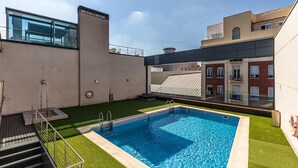 Una piscina en la azotea con barandilla de cristal, una pequeña escalera y vistas de edificios cercanos.