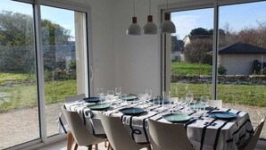 A dining area with a table set for six, large windows, and a view of a garden and house. - Le Gîte de la Grange de Dîme. Gite Tout Confort à 1 km du Phare de Gatteville (Gatteville-le-Phare)