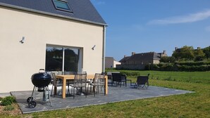 A house with a patio area featuring a barbecue grill, a dining table with chairs, and a large window. - Le Gîte de la Grange de Dîme. Gite Tout Confort à 1 km du Phare de Gatteville (Gatteville-le-Phare)