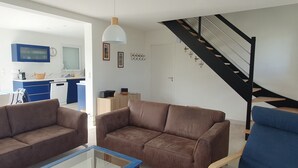 A modern living room with brown sofas, a glass coffee table, and a blue armchair. A staircase with black railings leads to an upper floor. - Le Gîte de la Grange de Dîme. Gite Tout Confort à 1 km du Phare de Gatteville (Gatteville-le-Phare)