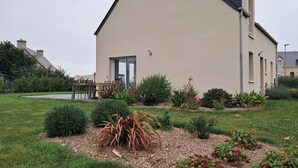 A modern house with a large front yard, a dining area with a table and chairs, and a variety of shrubs and plants. - Le Gîte de la Grange de Dîme. Gite Tout Confort à 1 km du Phare de Gatteville (Gatteville-le-Phare)