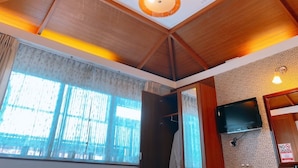 Une chambre d’hôtel avec un plafond haut, des poutres en bois, une grande fenêtre avec des rideaux, un téléviseur à écran plat fixé au mur et un panneau indiquant « interdiction de fumer ».