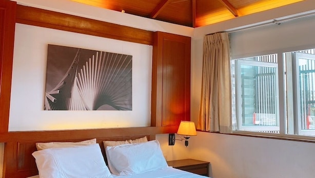 Une chambre d’hôtel avec un plafond en bois, un lit avec du linge de maison blanc, une œuvre d’art accrochée au mur, une lampe de chevet et une fenêtre avec des rideaux.