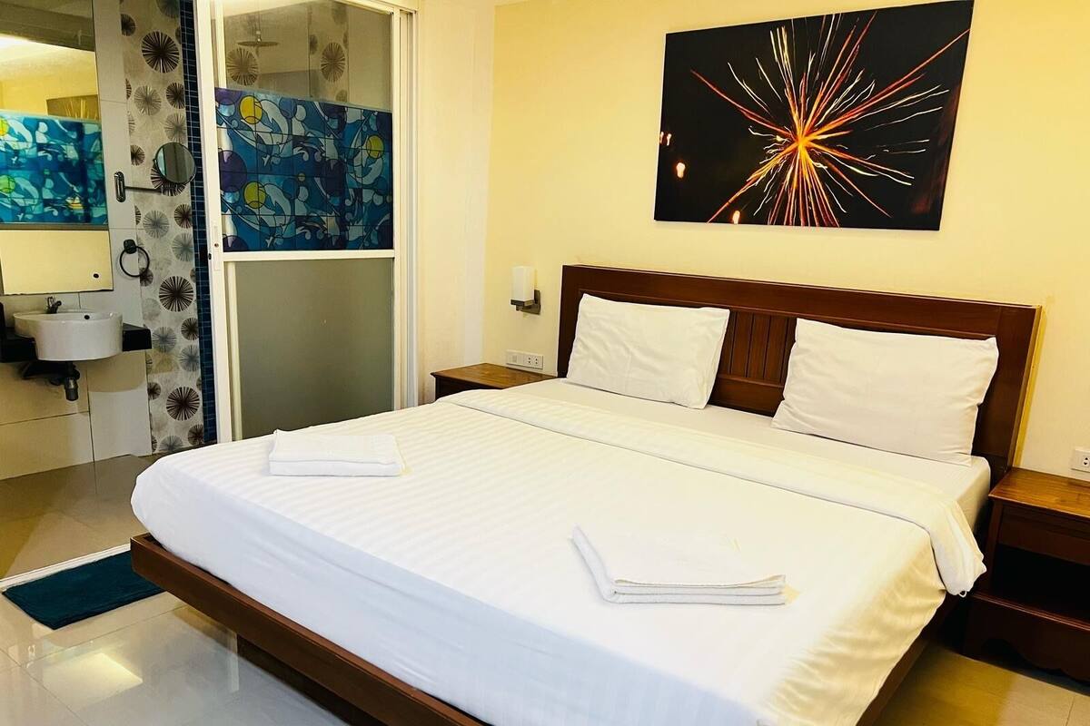 Une chambre d’hôtel avec un grand lit, une table de chevet, une commode, une salle de bain avec un lavabo et un miroir, et une peinture abstraite aux couleurs vives accrochée au mur.