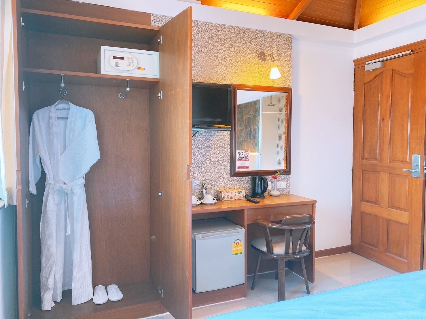 Een slaapkamer met een houten garderobe, een bureau met spiegel, een kleine koelkast en een stoel.