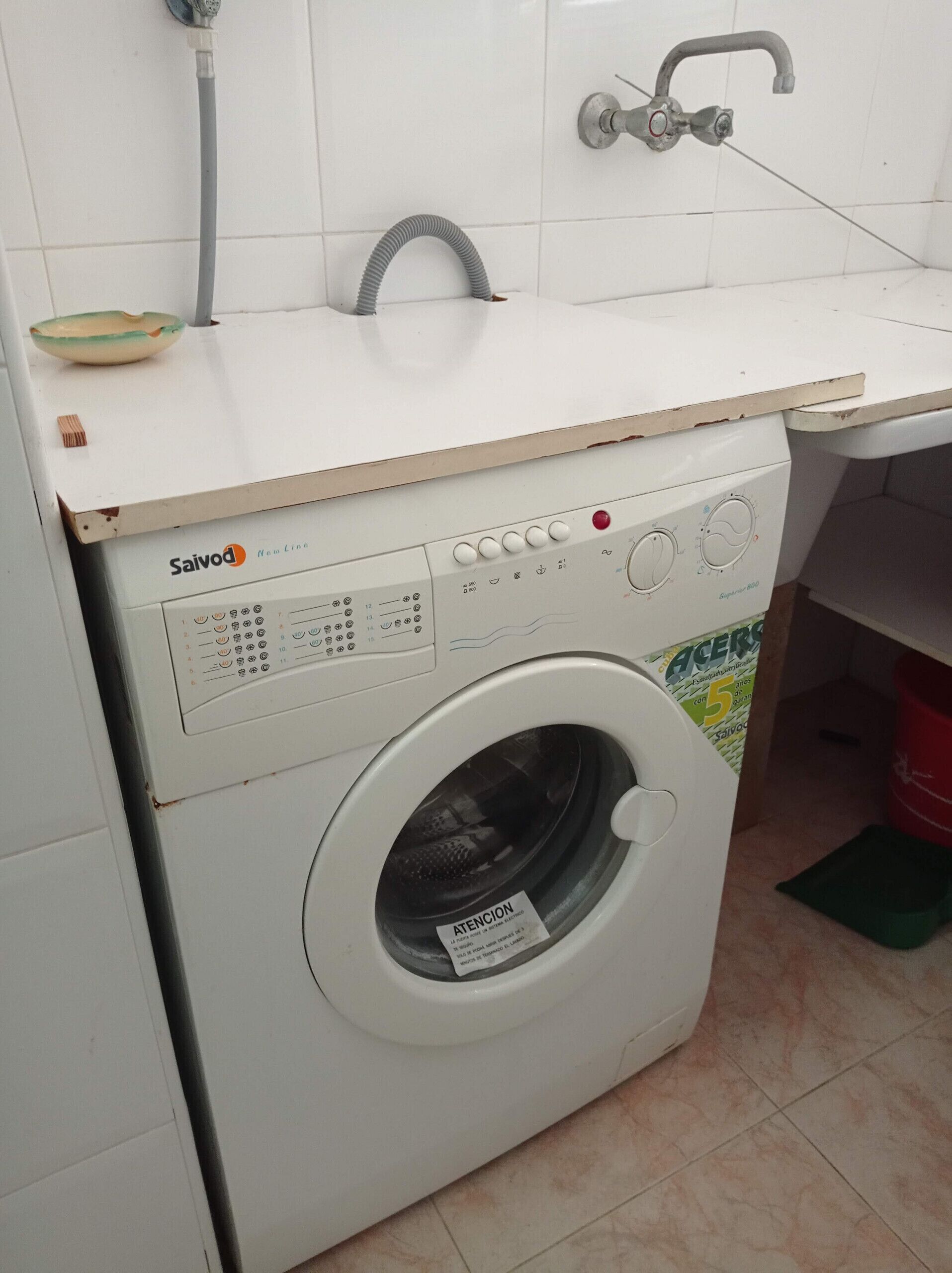 A front-loading washing machine with a control panel, a water inlet hose, and a tiled wall.