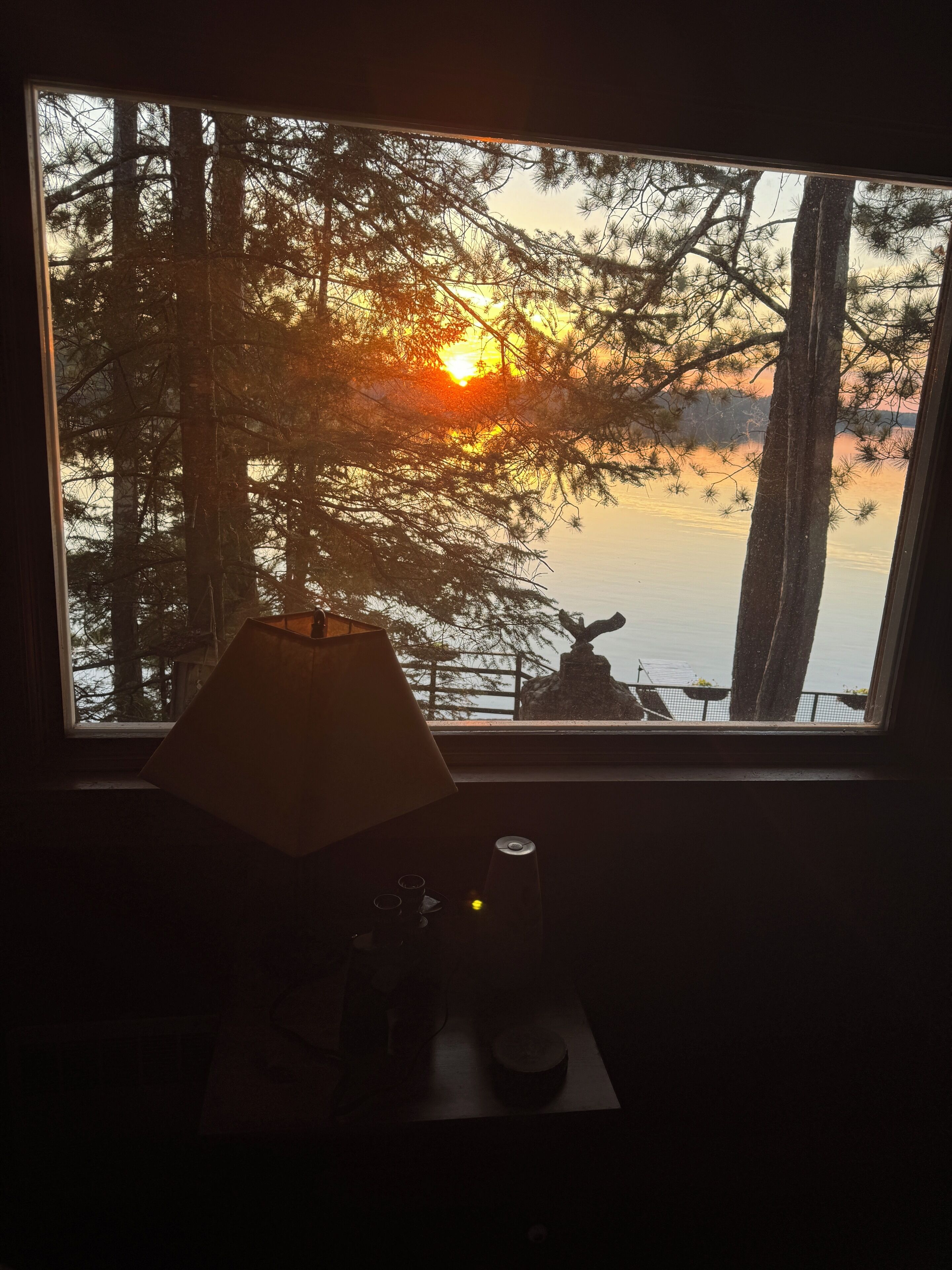 A room with a view of a sunset over a body of water, a lamp, and a small table with a cup and saucer.