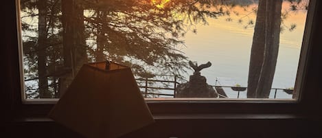 A room with a view of a sunset over a body of water, a lamp, and a small table with a cup and saucer.