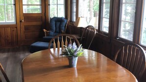 A wooden dining room with a round table, chairs, a chandelier, and large windows with a view of trees.
