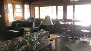 A cozy living room with a sofa, chairs, a table, and a lamp. There are large windows allowing natural light.