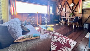 Une pièce chaleureuse avec un plafond en bois, un canapé agrémenté de coussins à motifs, une petite table avec des chaises et une grande fenêtre laissant entrer la lumière naturelle.