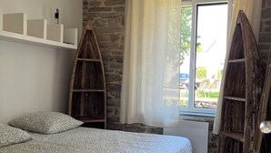 A bedroom with a bed, a stone wall, a window with curtains, and a hanging light fixture. - Maison Bretonne Typique Plage des Oiseaux à Fouesnant Beg Meil (Fouesnant)
