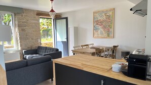 A compact living space with a stone wall, a grey sofa, a wooden dining table, and a kitchen area with a coffee machine. - Maison Bretonne Typique Plage des Oiseaux à Fouesnant Beg Meil (Fouesnant)
