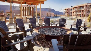 An outdoor seating area with Adirondack chairs and a fire pit, surrounded by buildings with balconies and a mountain view in the background.