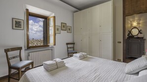 A bedroom with a large bed, a wooden chair, a radiator, a window with open shutters, and a wardrobe. - Casa Martina, Piazza Delle Erbe, San Gimignano (San Gimignano)