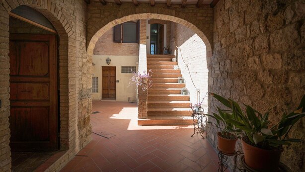 A stone-walled corridor with a wooden door, a staircase with a wrought iron railing, and potted plants. - Casa Martina, Piazza Delle Erbe, San Gimignano (San Gimignano)