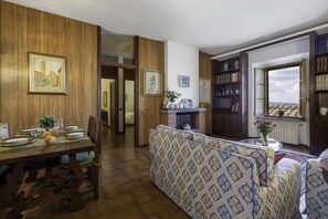 Un coin repas avec une table en bois dressée pour quatre personnes, un canapé recouvert d’un tissu à motifs et une cheminée agrémentée d’une étagère.