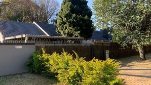 A fenced backyard with a paved path, a tall tree, and a building with a dark roof. - Saba Saba Guest House (Pretoria)